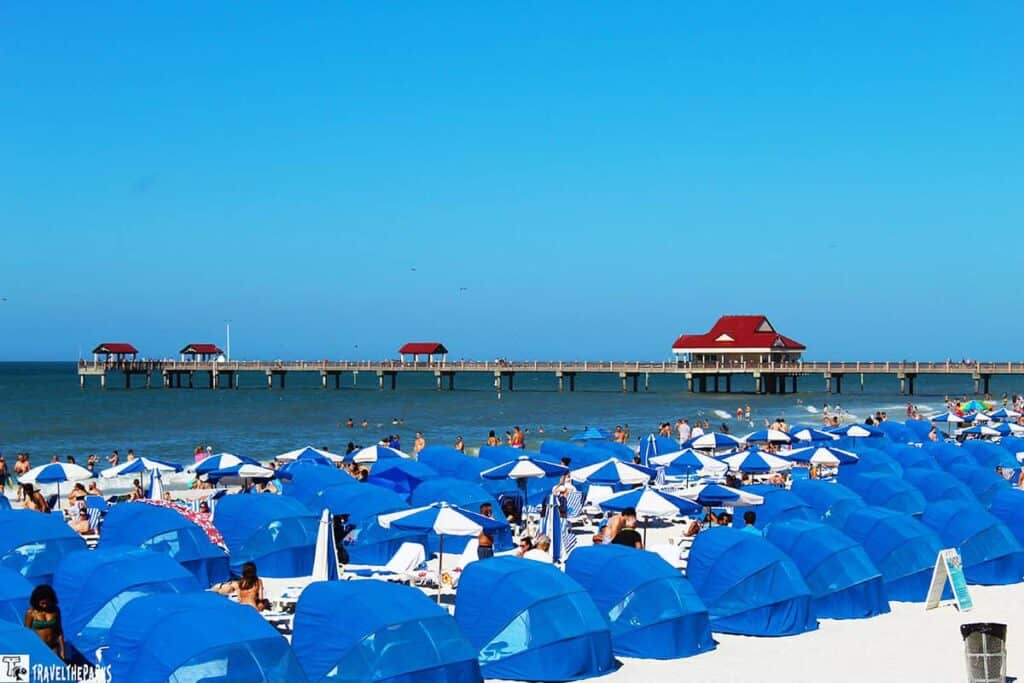 Crowded beach with blue umbrellas and a pier with red-roofed structures against a blue sky.

