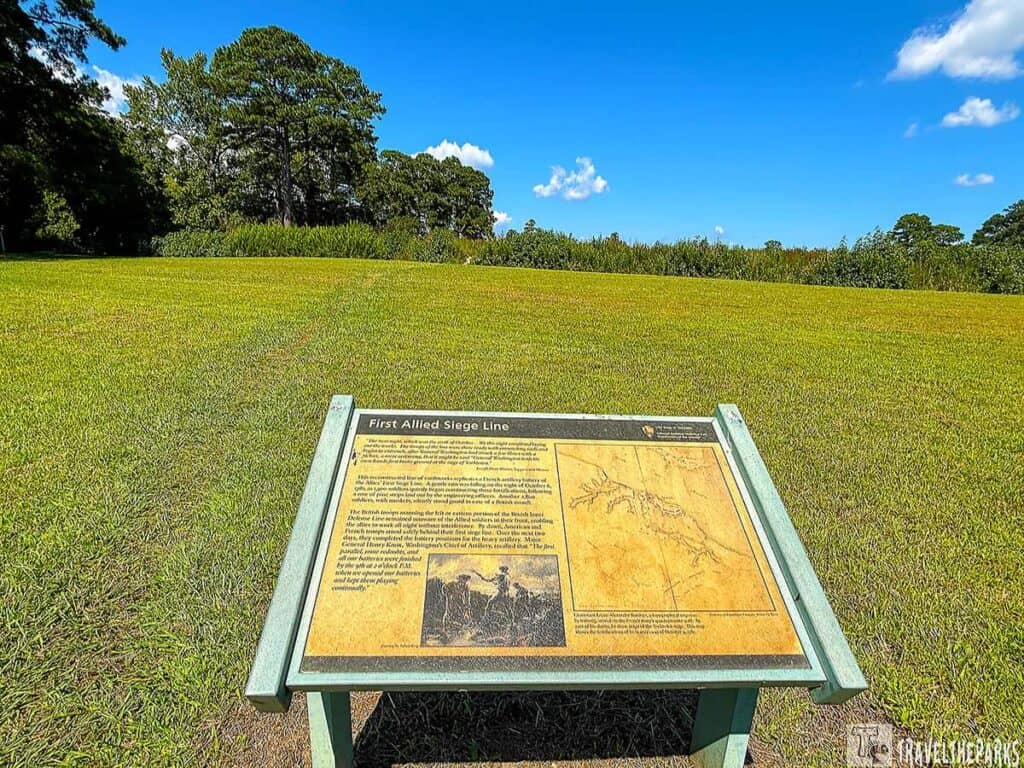 Historic site at Yorktown National Battlefield with a plaque titled "First Allied Siege Line" on a grassy field with trees and hedges in the background.