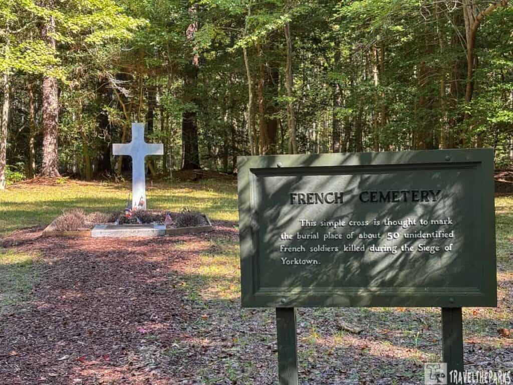A white cross and informational sign at the French Cemetery in a wooded area on the Yorktown National Battlefield drving tour.
