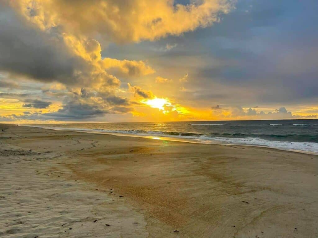 Beach at sunset with dramatic clouds and glowing sun near the horizon.