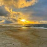Beach at sunset with dramatic clouds and glowing sun near the horizon.