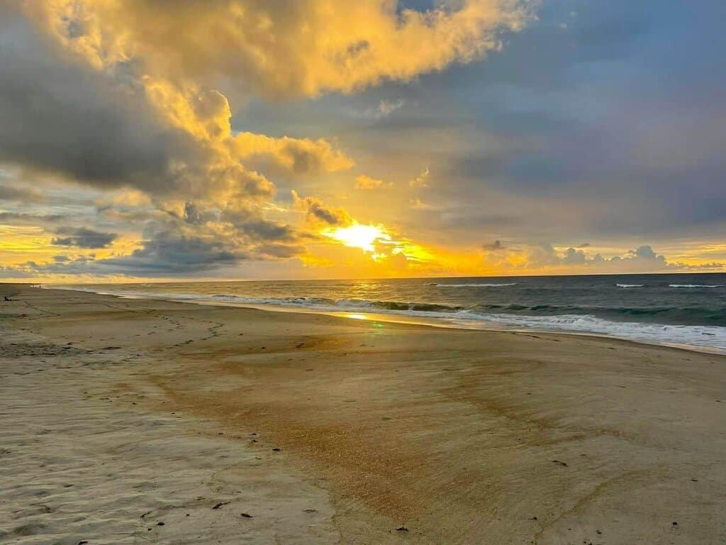 Beach at sunset with dramatic clouds and glowing sun near the horizon.