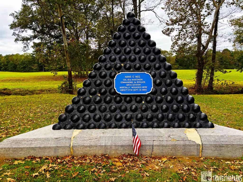 Monument to Colonel Hans Heg. Pyramid-shaped monument of black cannonballs with a blue plaque and small American flag at the base in a grassy, tree-lined park.


