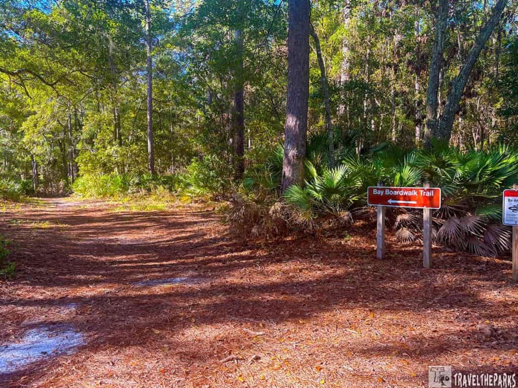 Trail entrance at Crooked River State Park with "Bay Boardwalk Trail" sign pointing left, surrounded by trees and greenery.