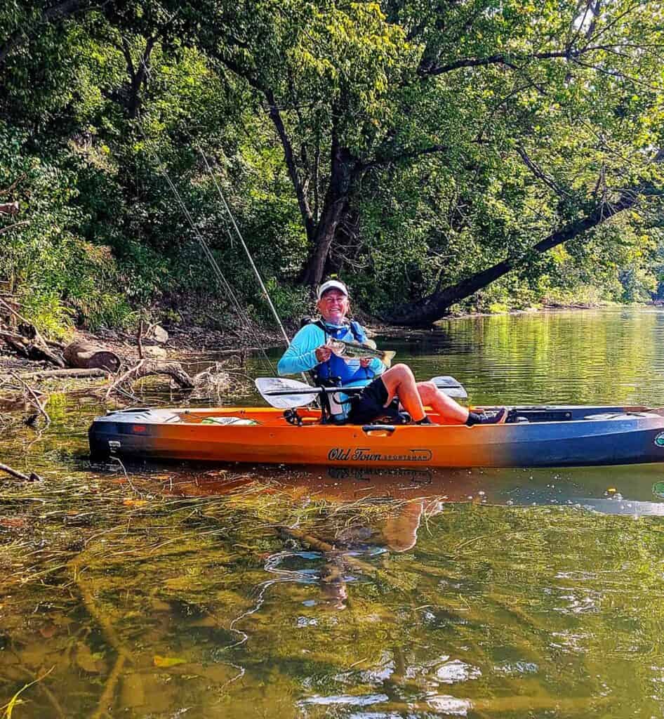 A person in an orange kayak holds a fish on a river with trees in the background.