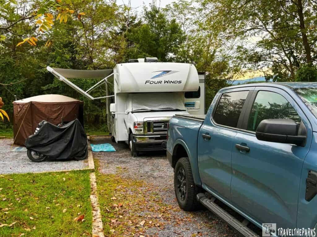 Campsite with a white RV, brown tent, covered motorcycle, and blue pickup truck in a wooded area.