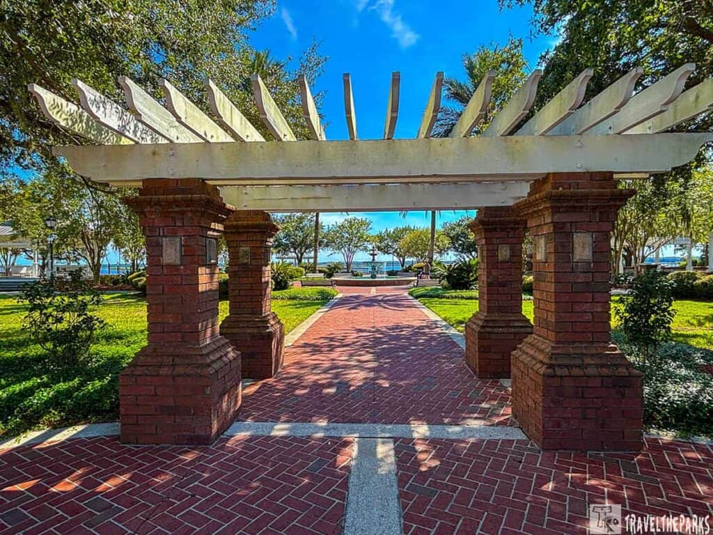 Brick-paved walkway under a pergola in a park leading to a view of trees and a waterfront.