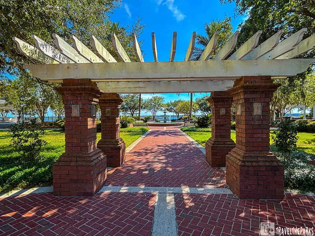 Brick-paved walkway under a pergola in a park leading to a view of trees and a waterfront.
