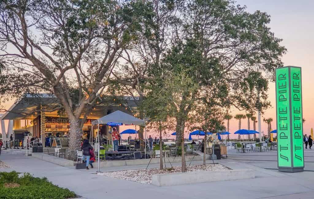 St. Pete Pier with a modern building, trees, seating areas, and a glowing sign at sunset.

