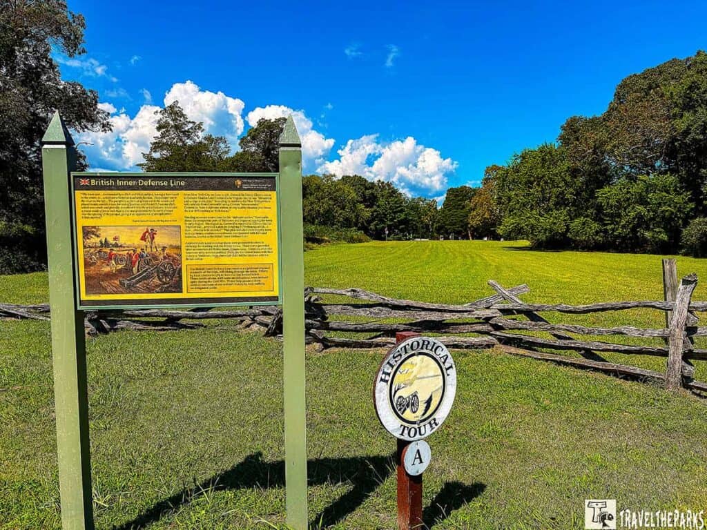 Informational sign and tour marker at Yorktown National Battlefield with a field and trees in the background under a blue sky.