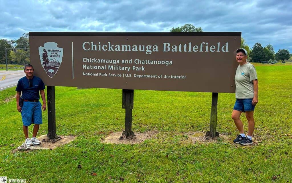 Two people stand beside a sign reading "Chickamauga Battlefield" with an overcast sky in the background.

