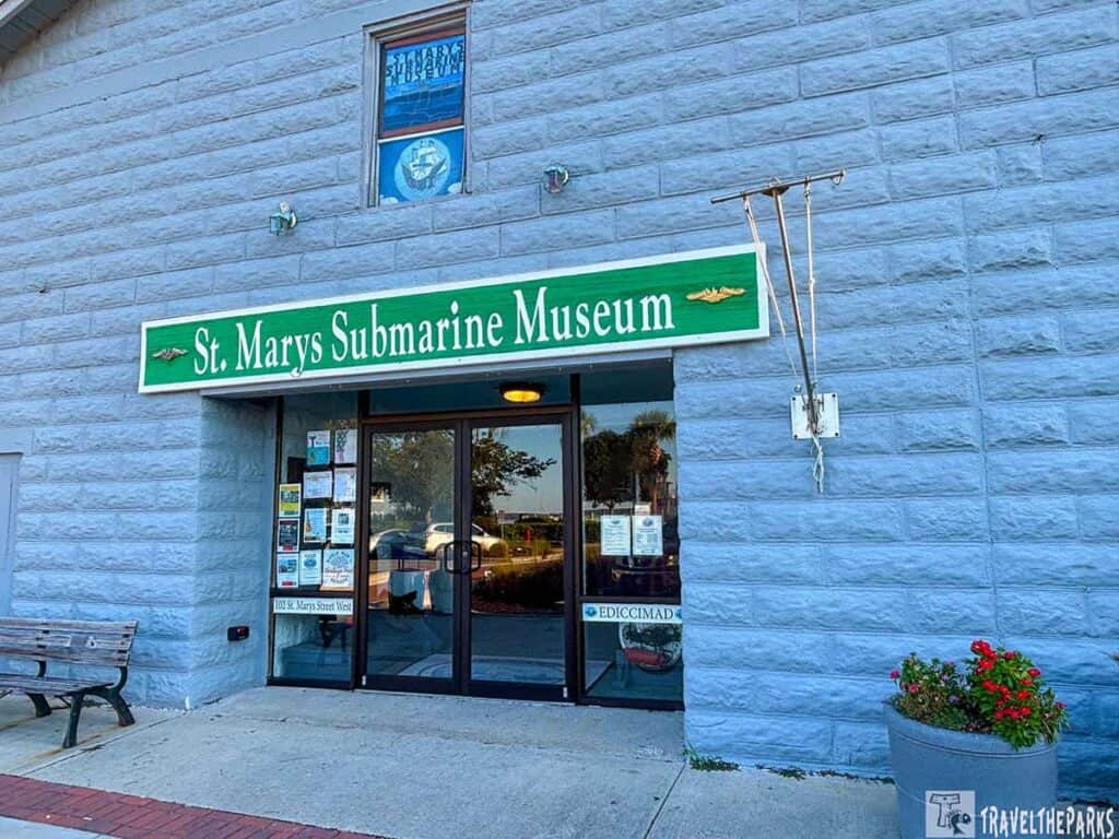 Entrance of St. Marys Submarine Museum with green signage above glass doors, a bench and a planter with flowers.