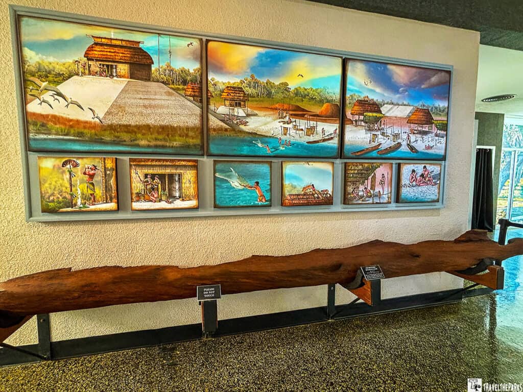 Wall-mounted paintings at Crystal River Archaeological State Park depicting indigenous life and structures, with a long wooden dugout canoe beneath.