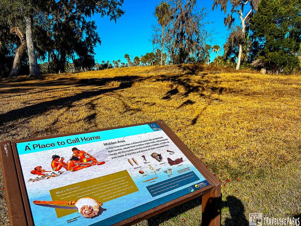 Informational sign about Midden Mound J-K with grassy field and oak trees in the background.


