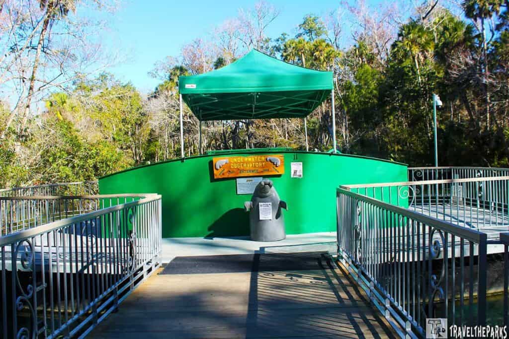 Green canopy covers a circular platform labeled "Underwater Observatory" with a manatee statue in front. Surrounded by lush trees and railings.