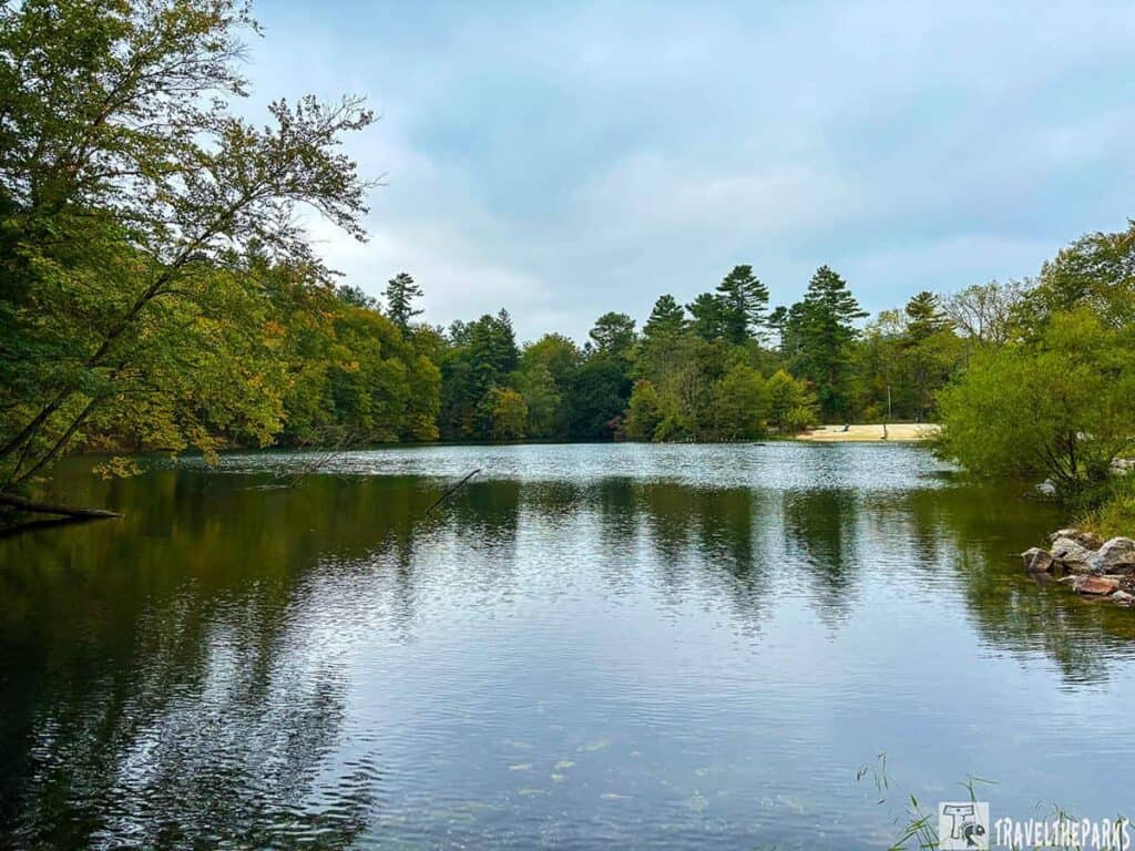 A peaceful lake surrounded by green and autumn-touched trees, with a sandy beach on the right.

