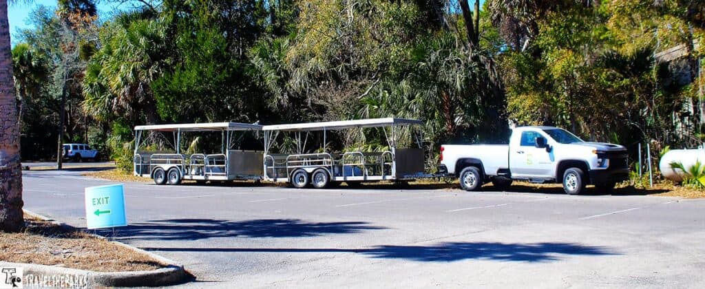 A white pickup truck with an attached covered trailer parked in an empty lot, surrounded by lush green trees. A sign with "Exit" and an arrow points left.
