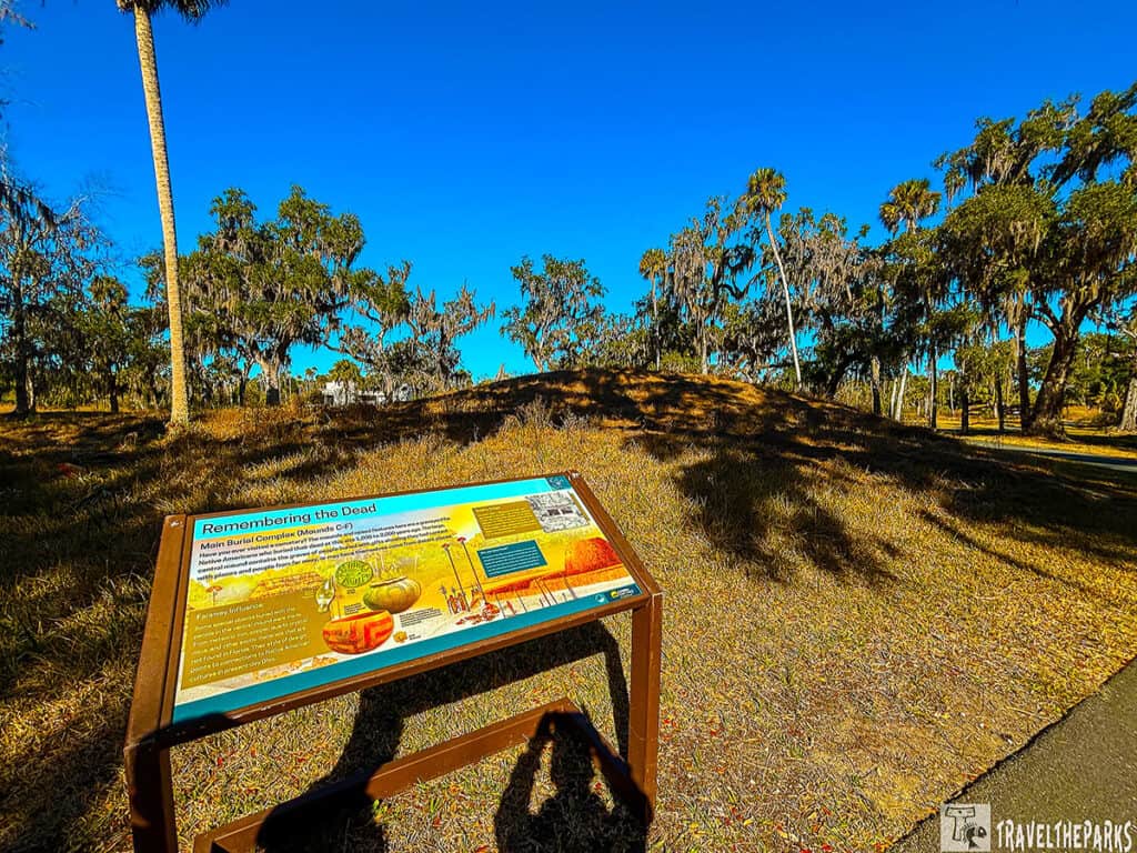 Informational sign Main-Burial-Complex-Mounds-C-F in front of a grassy mound with trees in the background at Crystal River Archaeological State Park.


