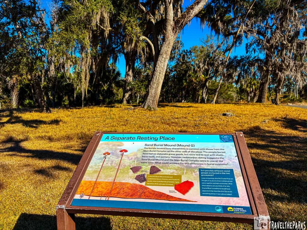 Informational sign about Sand Burial Mound G at Crystal River Archaeological State Park with trees and blue sky in the background.

