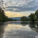 A calm river at sunrise or sunset with trees and mountain silhouettes at Shenandoah River State Park.