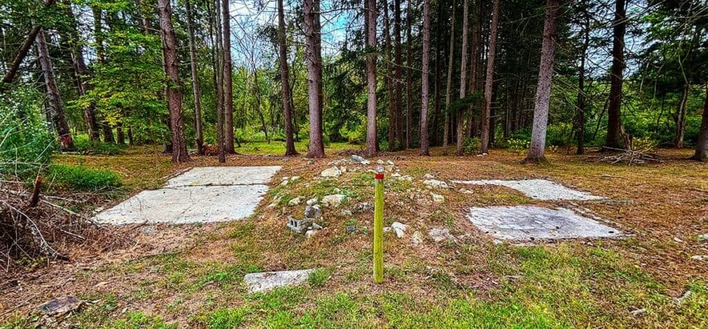 Camp Michaux Walking Tour-Stop 5: Concrete foundations of senior staff housing in a forest with a yellow post in the center. Self-guided walking tour of Camp Michaux