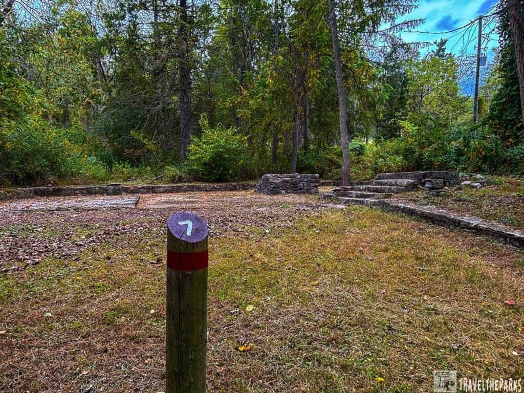POW Interrogation Center Building-Wooden post with number 7 in a wooded area with stone ruins and trees.