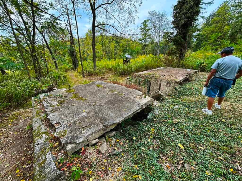 Bunker Hill farmhouse porch remains. Cracked concrete slabs in a wooded area with a person standing nearby.