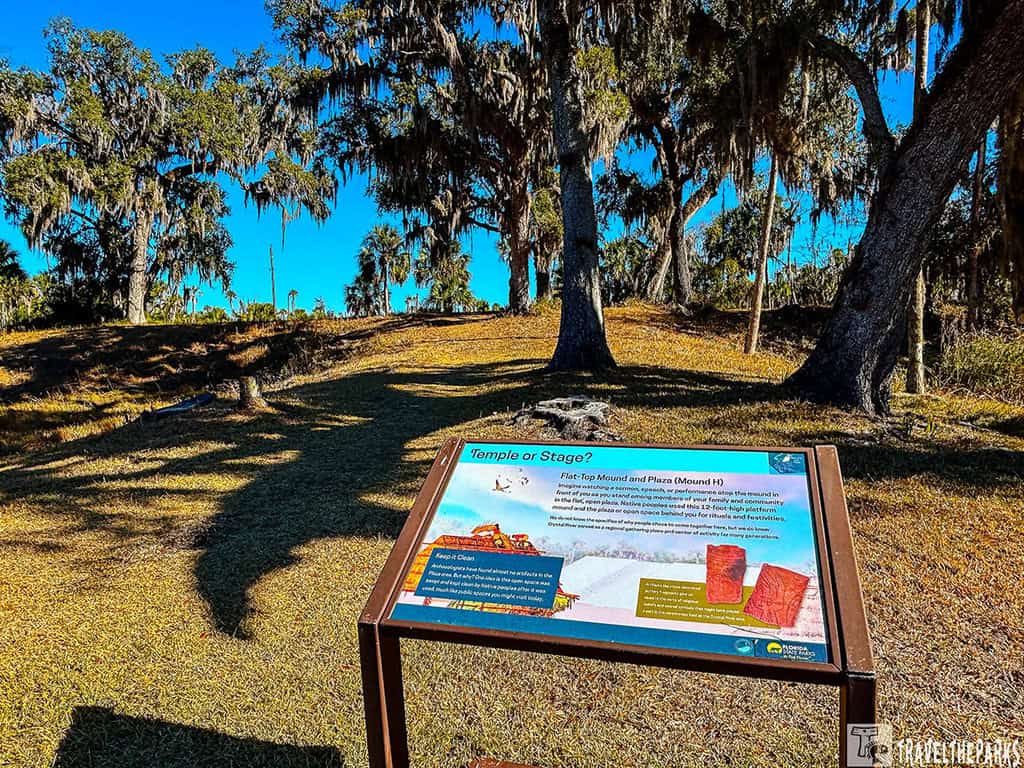 Informational sign Temple Mound H at the Crystal River Archaeological State Park with trees and a grassy hill in the background.