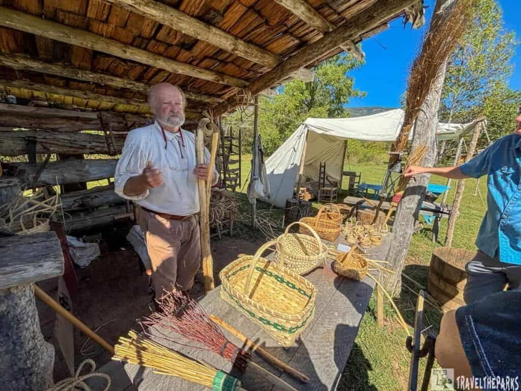A man in historical attire under a wooden shelter with handcrafted baskets at Martin Station, Wilderness Road State Park.