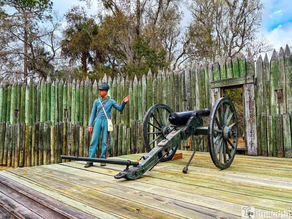 Person in historical military uniform next to a cannon in front of a wooden fort wall.