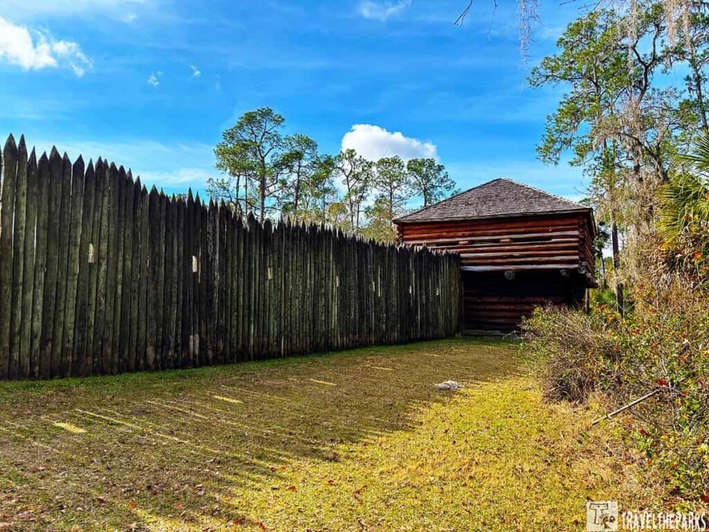 Wooden log fort with a tall fence at Fort Foster State Historic Site, surrounded by trees under a blue sky.