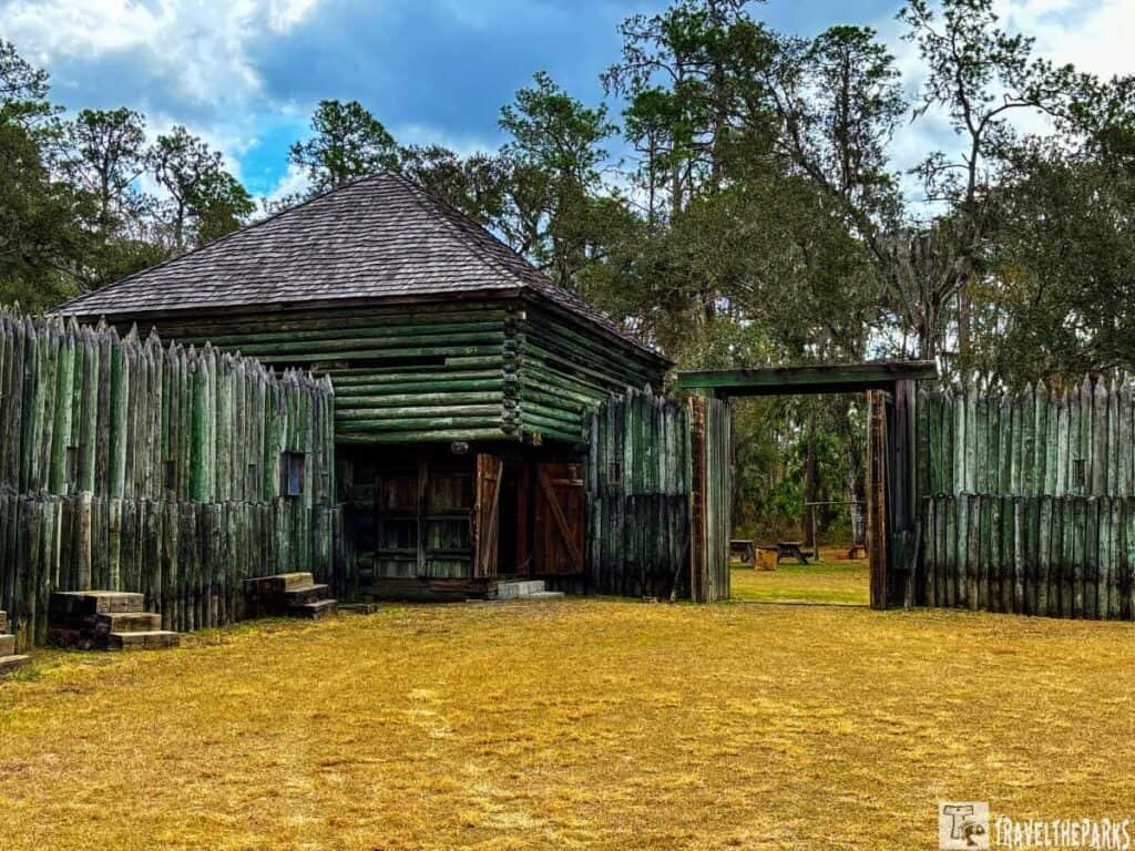 A historic fort surrounded by weathered wooden logs with an open gateway and trees in the background.