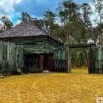 A historic fort surrounded by weathered wooden logs with an open gateway and trees in the background.