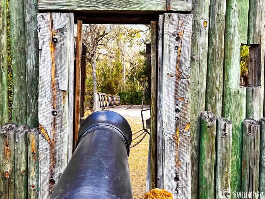 A black cannon pointing through a wooden fort Foster Histric Site entrance with weathered logs.