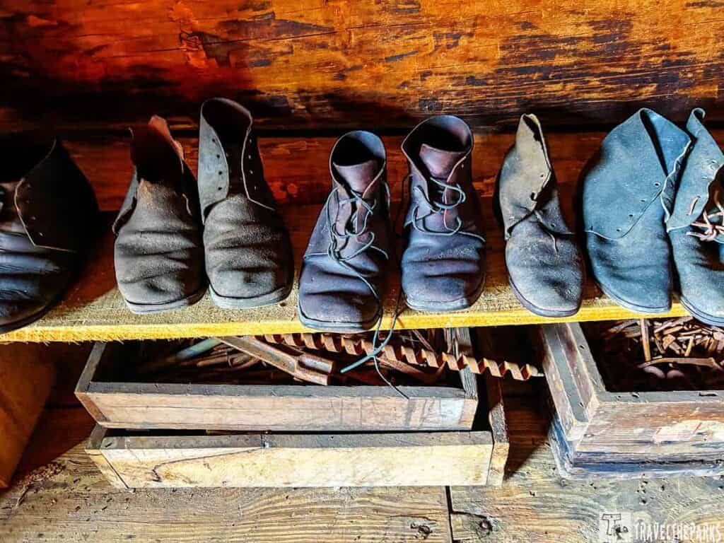 A row of worn black leather shoes on a wooden shelf, with crates of metal tools beneath.
