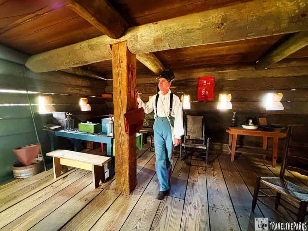 Interior of a rustic log cabin with a man in historical attire, wooden furniture, and vintage decor.