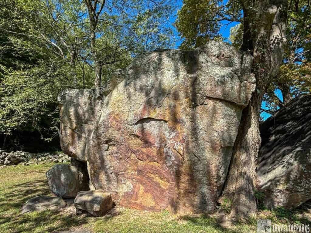 Large boulder with red and orange patches next to a tree in a grassy area.