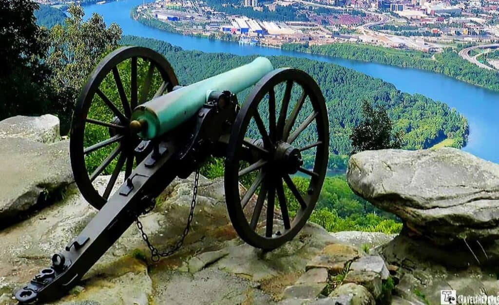 A historic cannon on a rocky ledge with a river and forested landscape in the background.