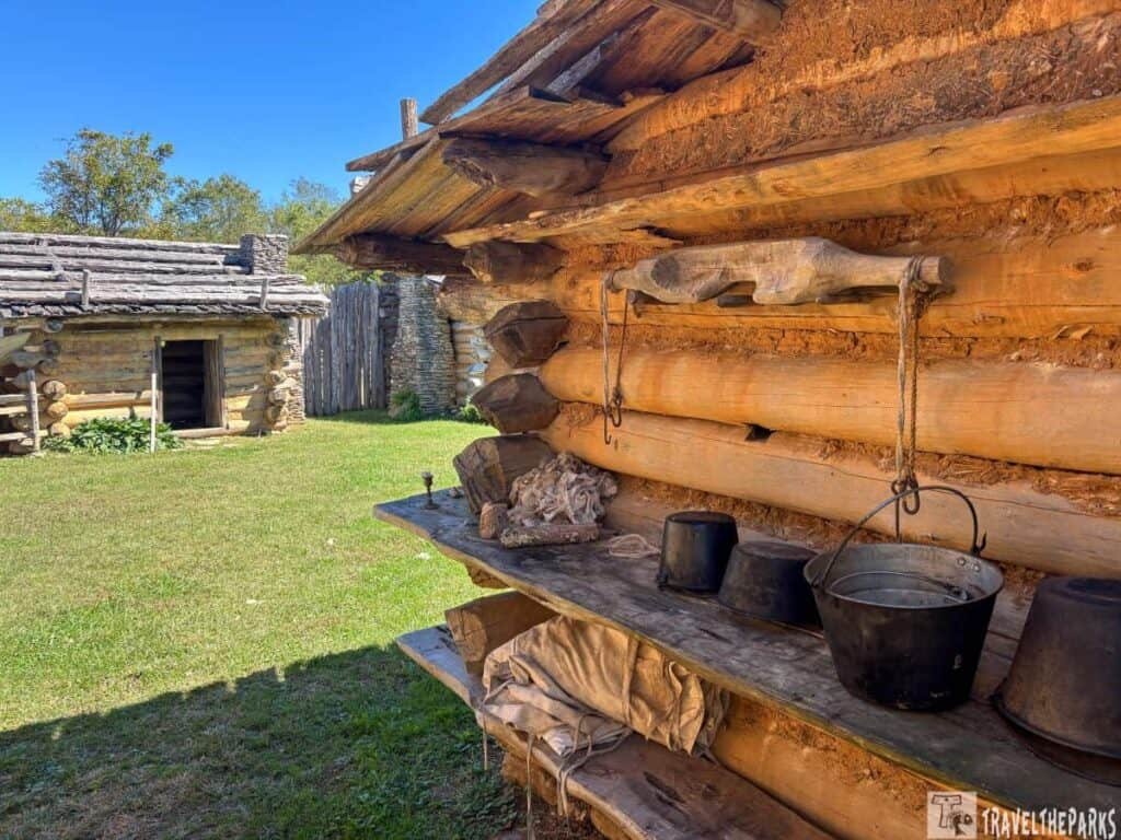 Log cabin at Martin Station with wooden shelves displaying rustic items and another cabin with a wooden door in the background.