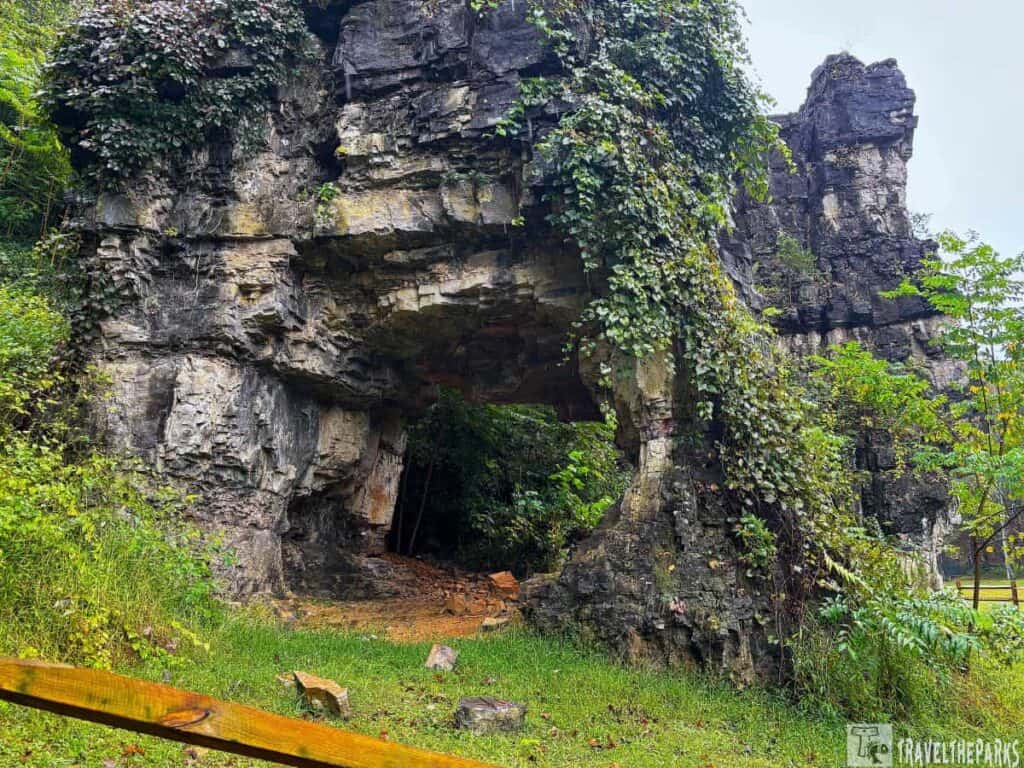 A natural rock archway with surrounding greenery and a wooden fence in the foreground.

