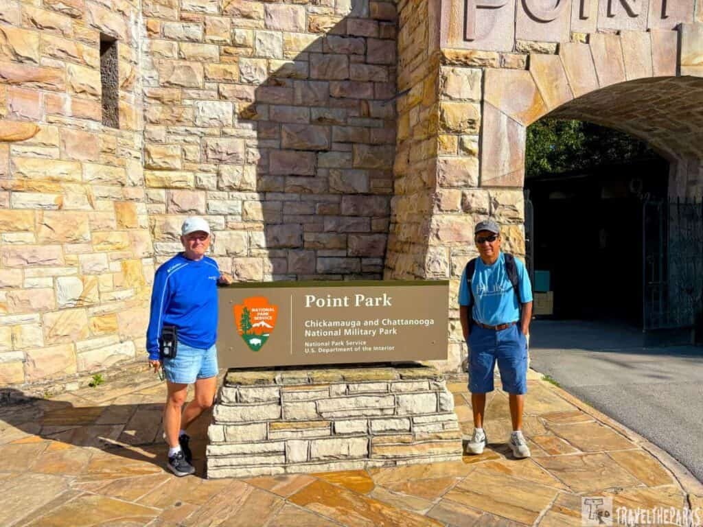 Two people stand by the Point Park sign at the entrance with a stone wall and archway backdrop.