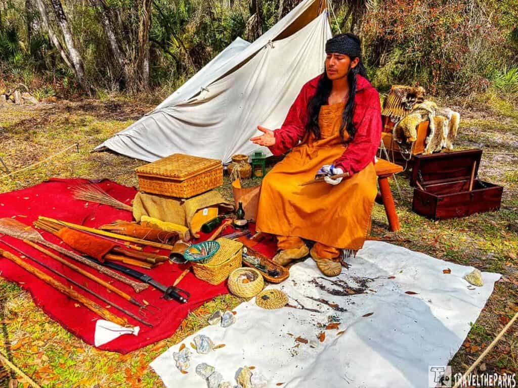 Person in historical Seminole indian attire seated outside a canvas tent with various historical artifacts displayed around them.