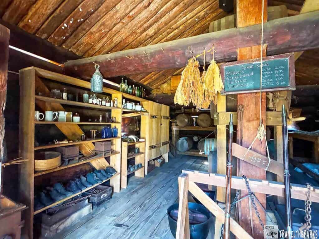 Interior of a rustic historical cabin with shelves, dried plants, rifles, and a chalkboard at Fort Foster State Historic Site.