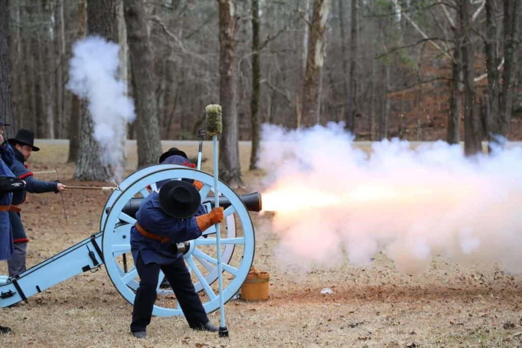 A historical reenactment of a cannon firing drill at Horseshoe Bend National Military Park, with three individuals in period attire operating a light blue cannon with smoke and flame visible.