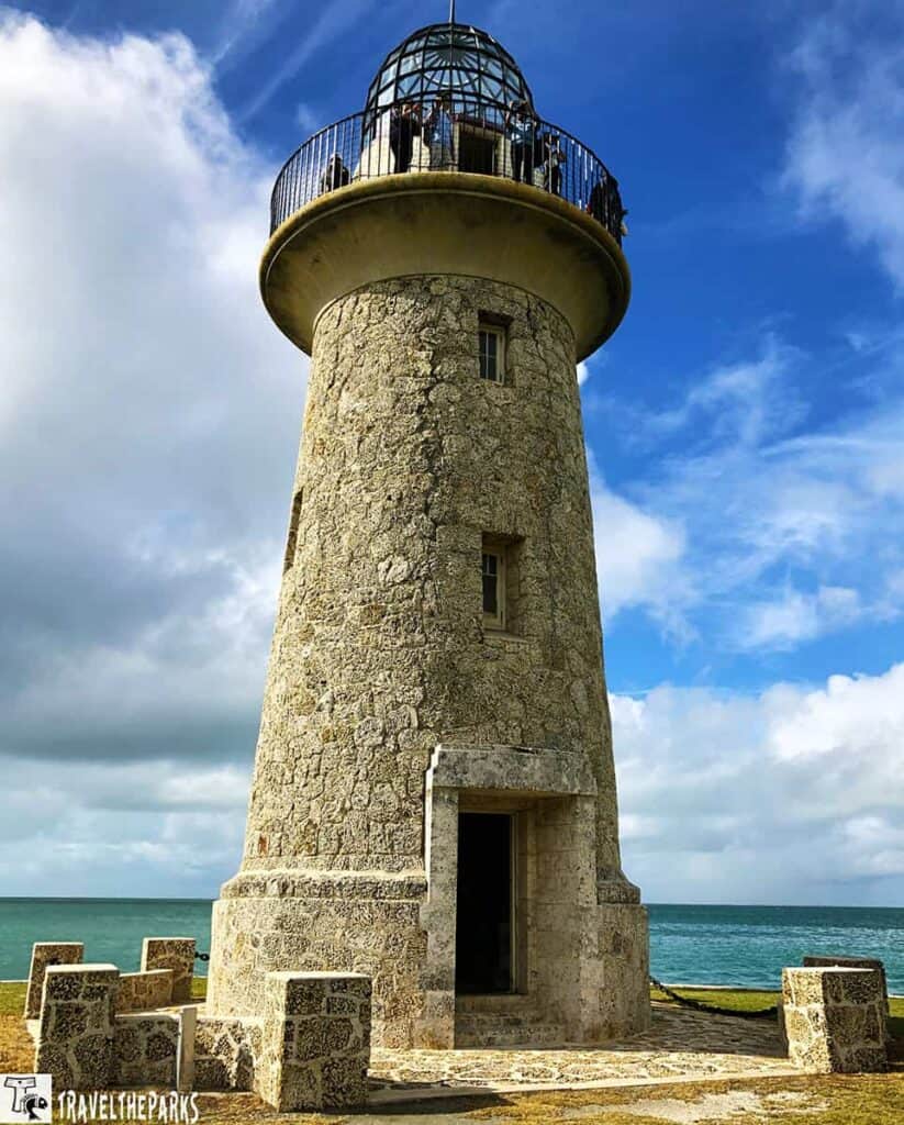 Boca Chita Lighthouse, a stone structure standing prominently against a backdrop of blue sky and ocean. The lighthouse features a cylindrical gray stone tower with a rough, textured surface.