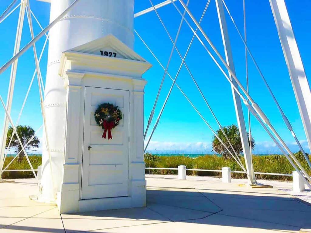 Base of Gasparilla Island Lighthouse with a door, wreath, and the year 1927; palm trees and ocean in the background.

