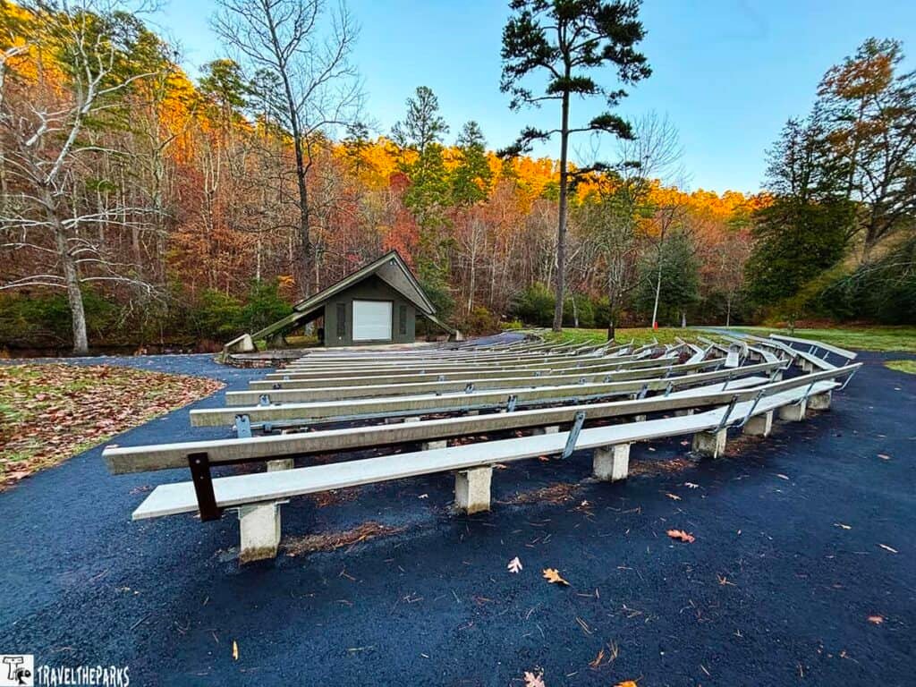 Outdoor amphitheater with wooden benches and a small structure, set in a forest with colorful autumn trees.