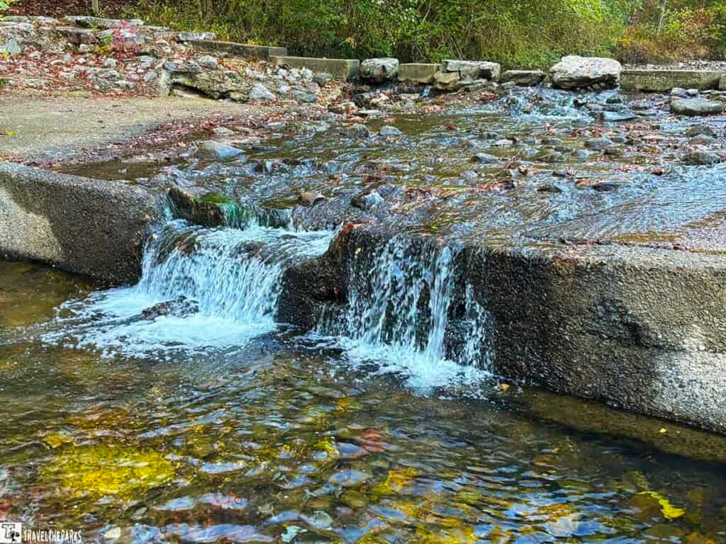 Small stream cascading over a concrete embankment with surrounding rocks and greenery.