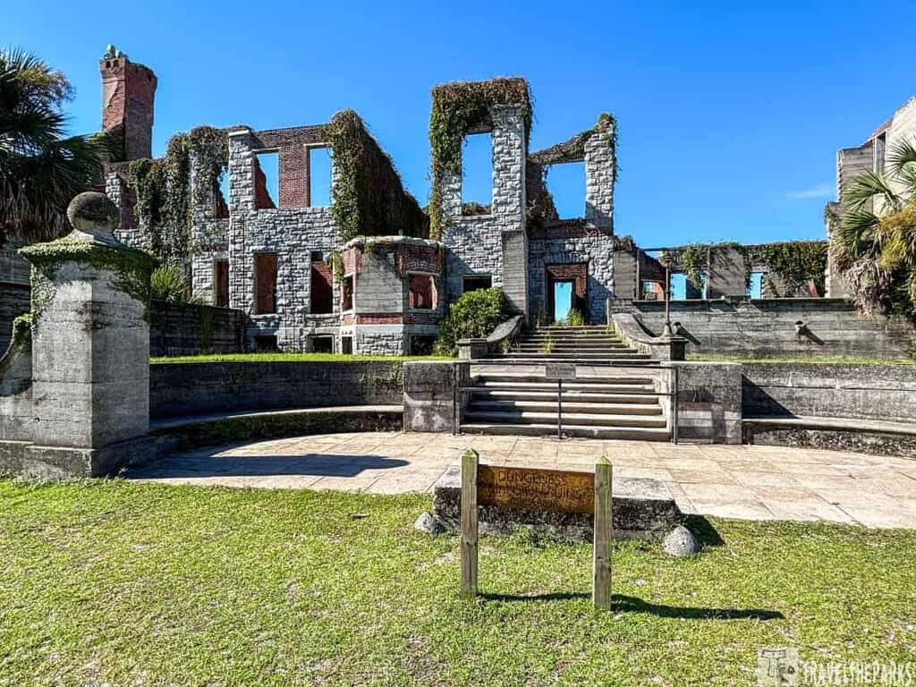 Dungeness Mansion: Stone ruins with tall windows and a brick chimney, partially covered in greenery, with a staircase leading up to them.