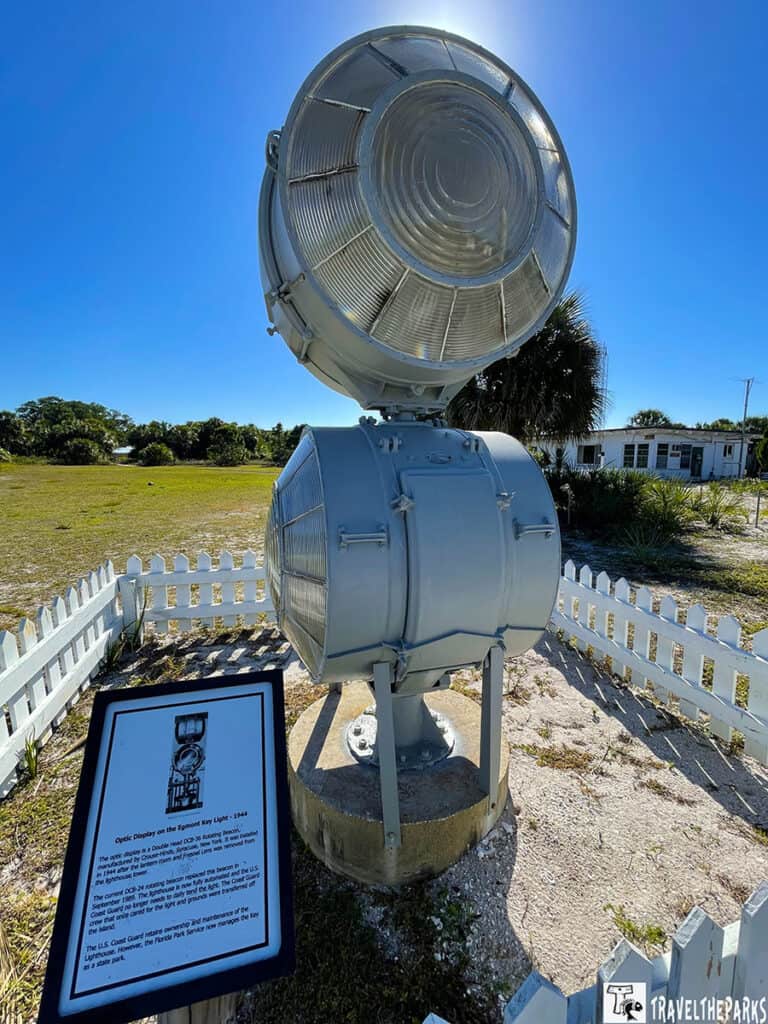  vintage lighthouse optic on display with a large glass lens, surrounded by a white picket fence, under a clear blue sky.

Transcribed Text:

"Optic Display on the Egmont Key Light - 1944

The optic display is a Double Head (2D) 36 Rotating Beehive Fresnel lens by Chance / Henry-Lepaute of Paris. It was used on the island as part of a lighthouse replacement lens system removed in 1984.

This current D2H optic was initially used as the beacon in the St. George's Reef Lighthouse, California from 1912-1971. The U.S. Coast Guard proudly incorporated this lens into the Egmont Light. It has one layer for the bright light and several beehives of lens glass on the exterior for equal diffusion of the light. The Guard continues as the keeper of the fire and the U.S. Park Service of the property.

The U.S. Coast Guard retains ownership and maintenance of the lighthouse. However, the Florida Park Service now manages the key as a state park."